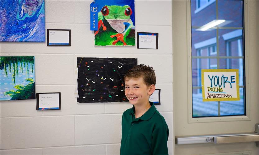 A boy wearing a green shirt smiles while standing beneath a drawing of a frog
